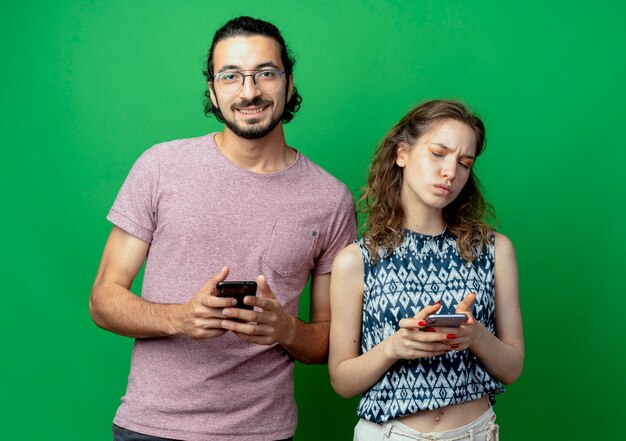 young beautiful couple man and women, man smiling holding smartphone standing next to his displeased girlfriend with cellphone over green wall