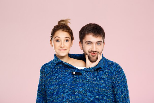 Young beautiful couple in one blue knitted sweater posing smiling having fun over light pink wall