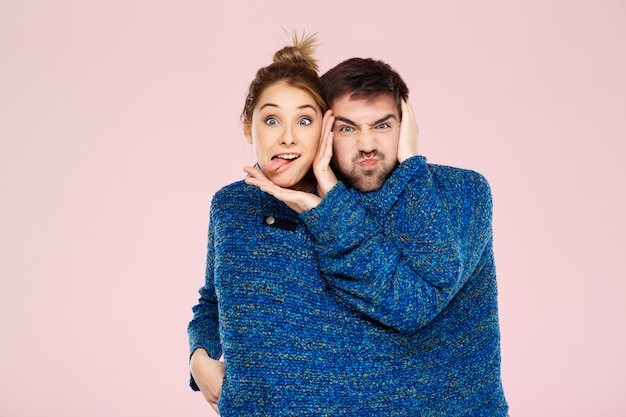 Free photo young beautiful couple in one blue knitted sweater posing smiling having fun over light pink wall