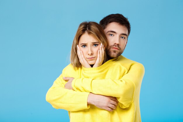 Young beautiful couple in one yellow sweater embracing smiling over blue wall