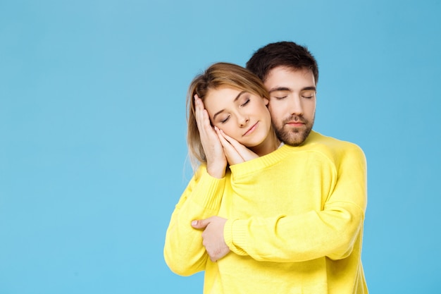 青い壁に笑みを浮かべて抱きしめる1つの黄色いセーターの若い美しいカップル