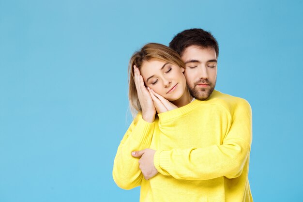 Young beautiful couple in one yellow sweater embracing smiling over blue wall