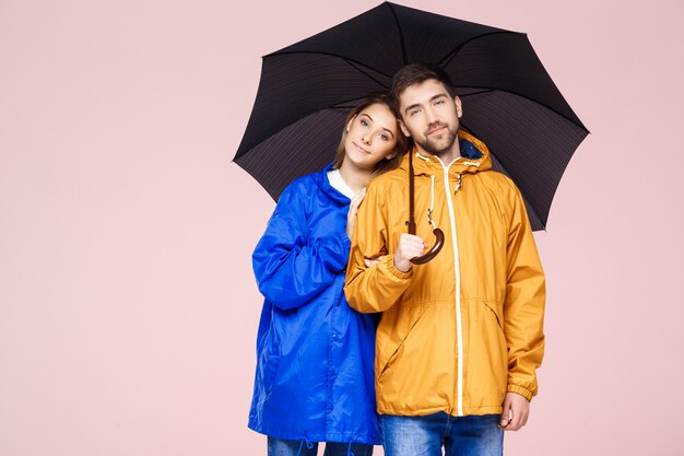 Young beautiful couple posing in rain coats holding umbrella over light pink wall