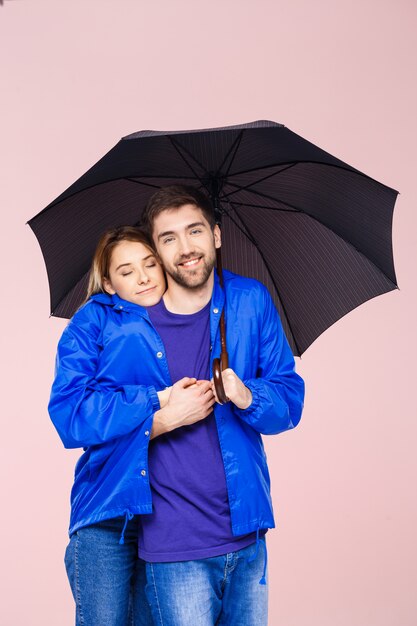 Young beautiful couple posing wearing one rain coat holding umbrella over light pink wall