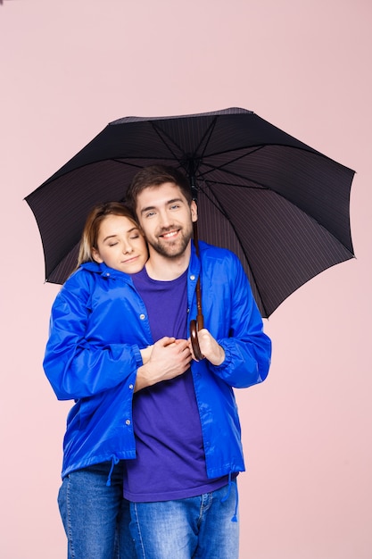 薄ピンクの壁の上に傘を保持している1つのレインコートを着てポーズ美しいカップル