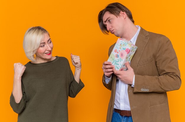 Young beautiful couple sad man giving a present to his lovely surprised girlfriend happy in love together celebrating international women's day standing over orange wall