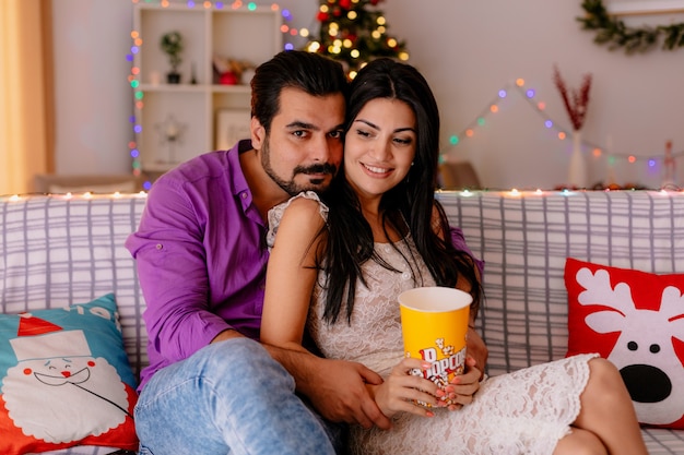 молодая и красивая пара сидит на диване мужчина и женщина с ведром попкорна вместе смотрят телевизор счастливы в любви в украшенной комнате с рождественской елкой на заднем плане