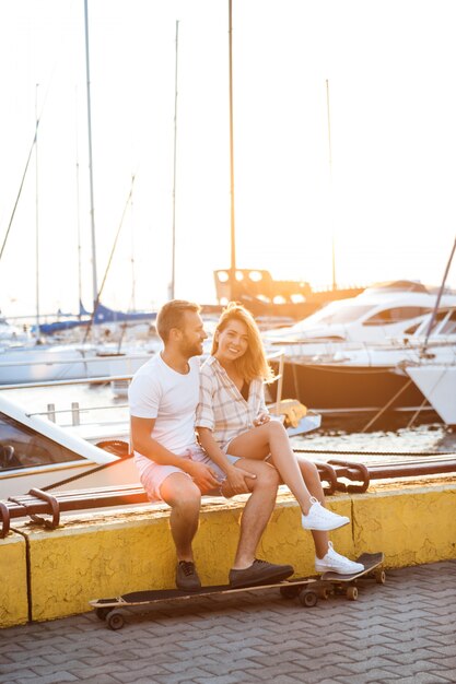 Young beautiful couple sitting at seaside, smiling, skateboarding.