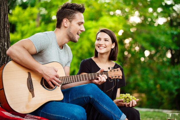 Young beautiful couple smiling, resting, relaxing on picnic in park. Man playing guitar. Copy space.