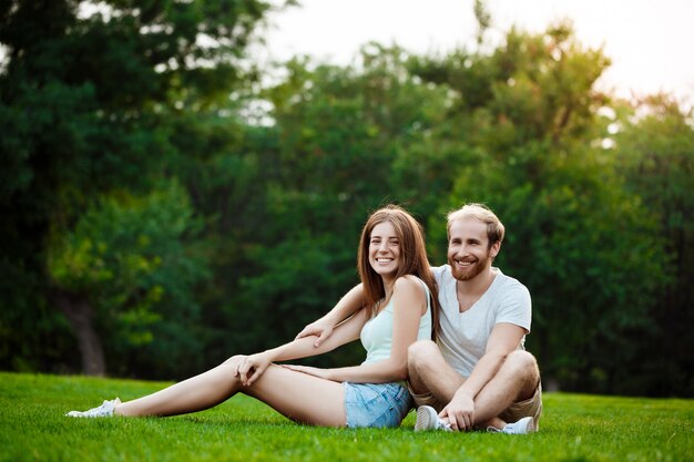 笑みを浮かべて、公園の芝生の上に座っている若い美しいカップル