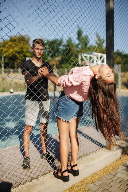 Young beautiful couple standing between mesh fence and holding hands of each other on basketball court