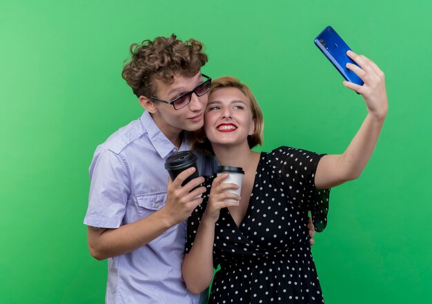 Young beautiful couple standing together holding coffee cups using mobile phone taking selfie smiling over green wall