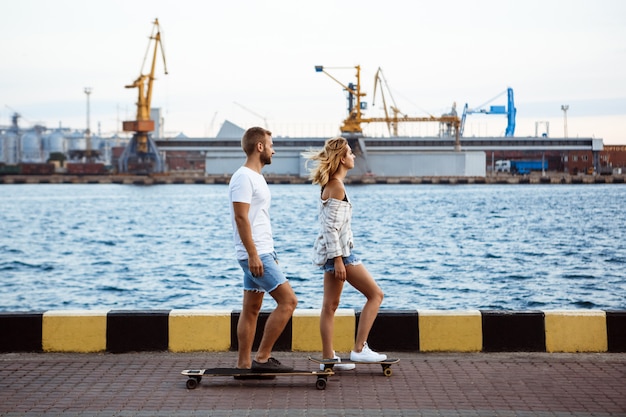 海辺を歩いて、笑って、スケートボードの美しいカップル。