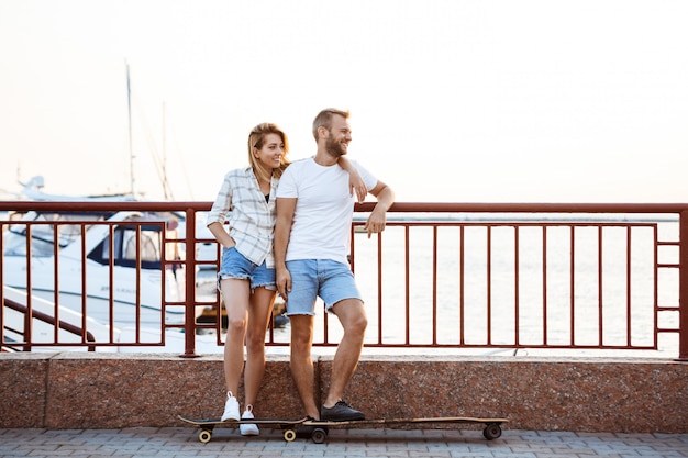 Young beautiful couple walking at seaside, smiling, skateboarding.