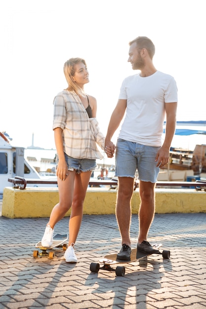 Young beautiful couple walking at seaside, smiling, skateboarding.
