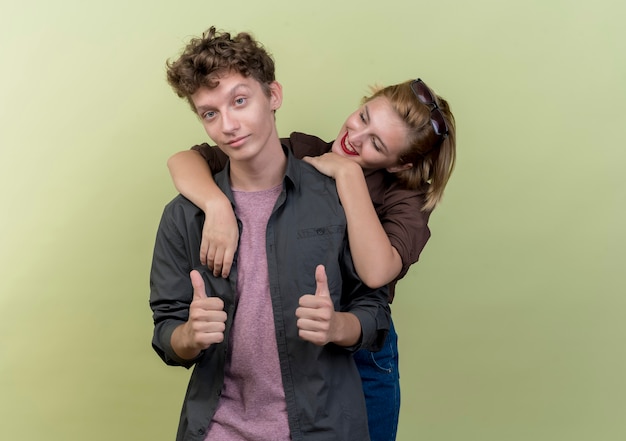 Young beautiful couple wearing casual clothes boy and girl happy in love girl hugging her boyfriend while he showing thumbs up over light