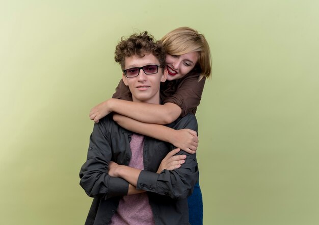 Young beautiful couple wearing casual clothes having fun together boy carrying his girlfriend piggyback standing over light wall