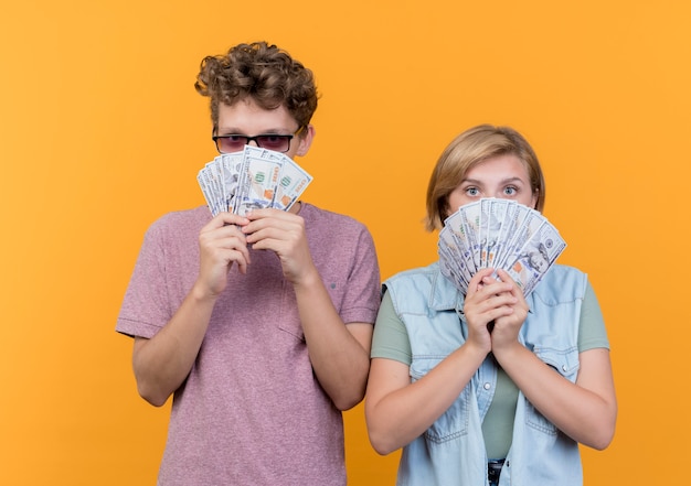 Бесплатное фото Молодая красивая пара в повседневной одежде мужчина и женщина, показывающие деньги, выглядят удивленными над оранжевым