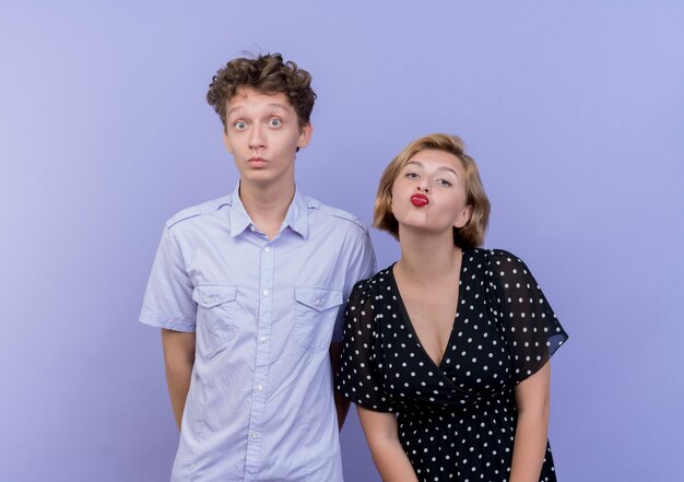 Young beautiful couple woman blowing a kiss while her boyfriend  confused and surprised standing over blue wall