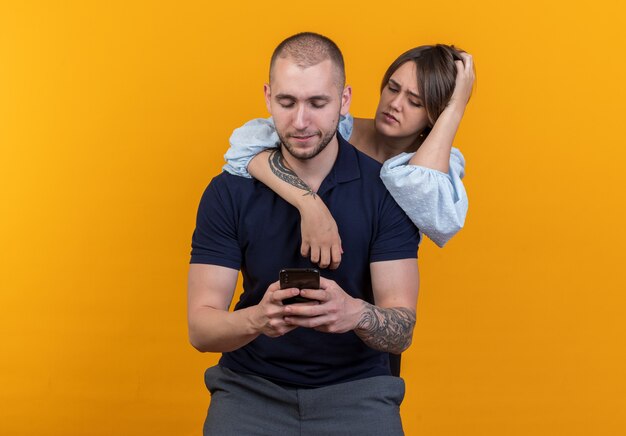 Young beautiful couple woman standing behind her boyfriend looking suspiciously at his smartphone