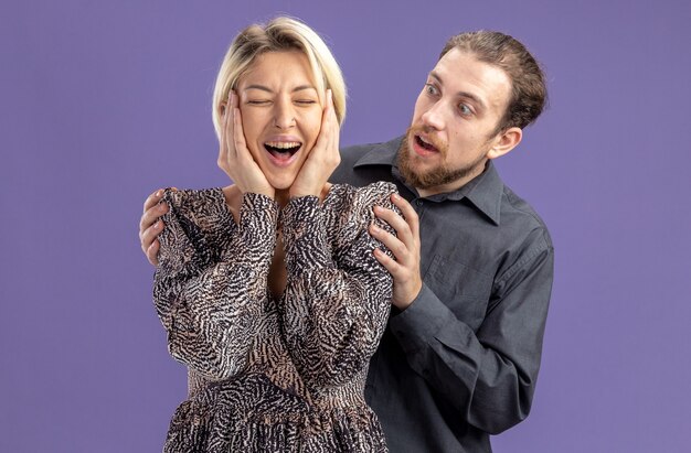 young beautiful couplesurprised man looking at his crazy happy girlfriend valentines day concept standing over purple wall