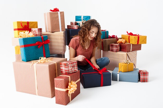 Young beautiful curly girl sitting on floor among gift boxes opening one of them Isolated