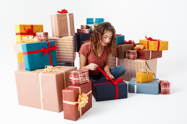Молодая красивая кудрявая девушка сидит на полу среди подарочных коробок, открывая один из них изолированных