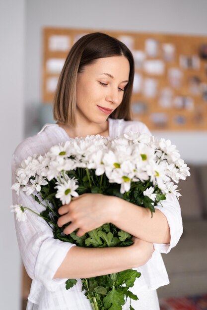 흰색 벽 배경에 흰색 신선한 꽃 꽃다발을 들고 있는 젊고 귀엽고 사랑스러운 웃는 여자