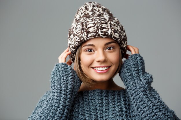 Young beautiful fair-haired woman in knited hat and sweater smiling on grey.