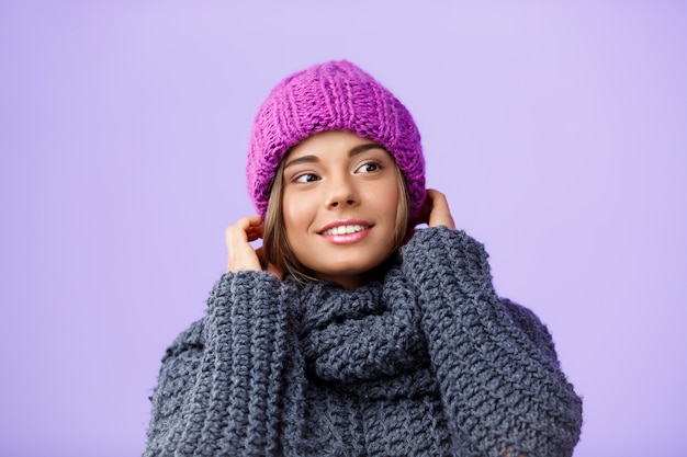 紫の側を見て笑っているニット帽子とセーターの若い美しい金髪の女性。