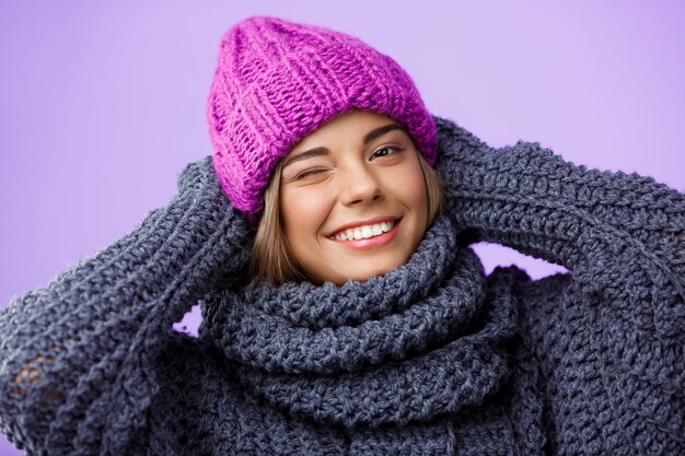 Young beautiful fair-haired woman in knited hat and sweater smiling winking on violet.