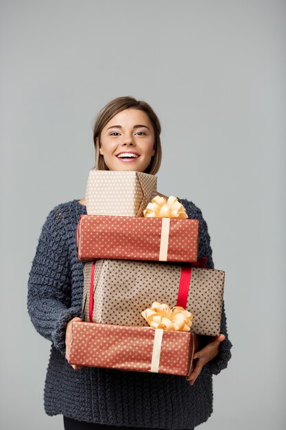 Young beautiful fair-haired woman in knited sweater smiling holding gift boxes on grey.