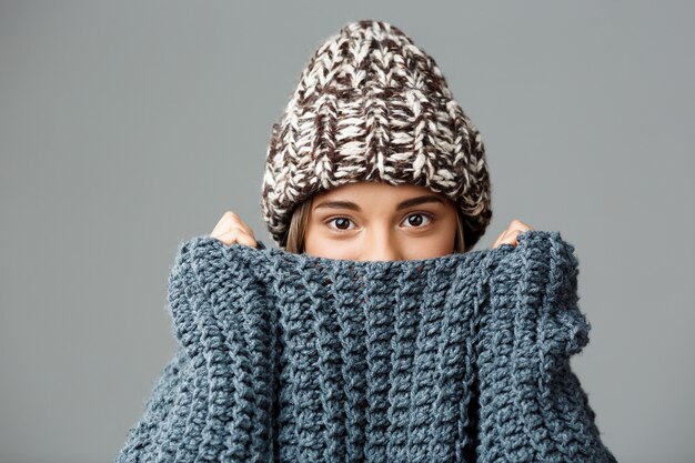 Young beautiful fair-haired woman wearing knited hat and sweater hiding her face in collar on grey. Copy space.
