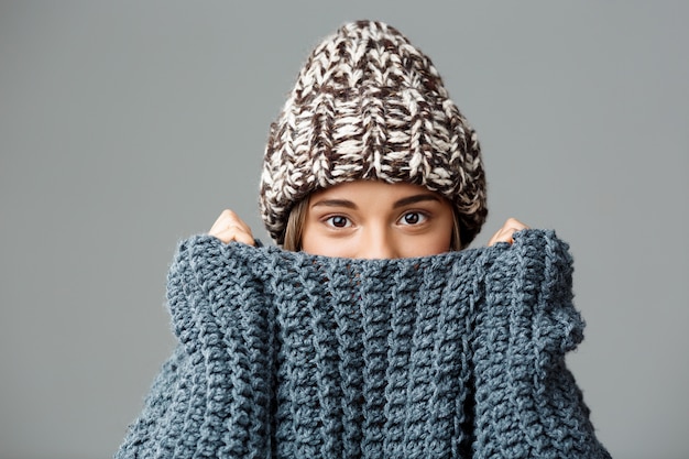 Молодая красивая белокурая женщина в вязаной шапке и свитер, скрывая ее лицо в воротник на серый. Копировать пространство