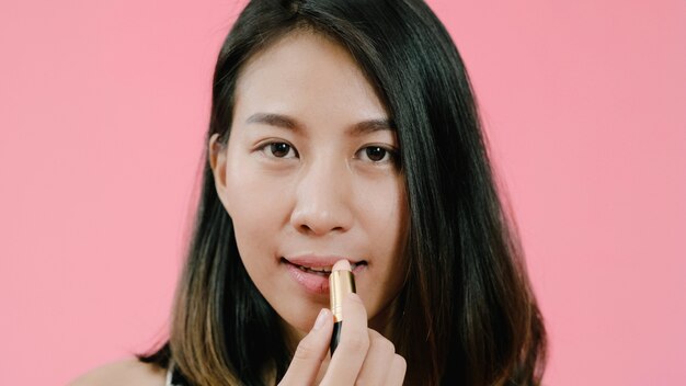 Молодая красивая модная азиатская женщина прикладывая состав губ с косметической щеткой в вскользь одежде над розовой съемкой студии предпосылки.