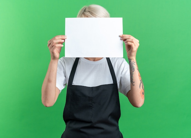 Young beautiful female barber in uniform covered face with papper isolated on green background