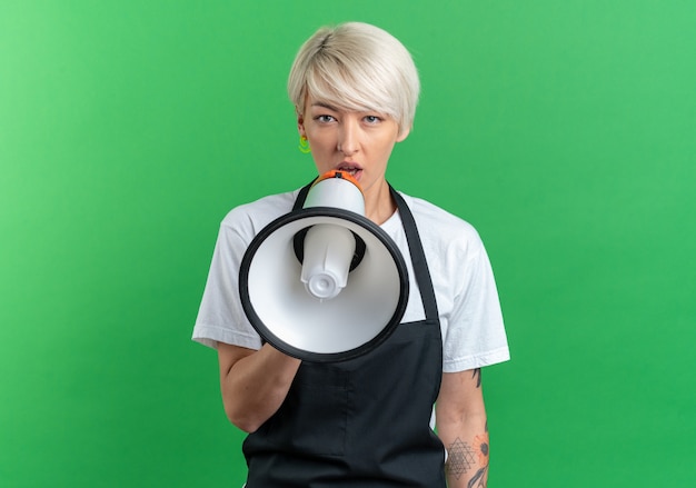 Young beautiful female barber in uniform speaks on loudspeaker isolated on green wall