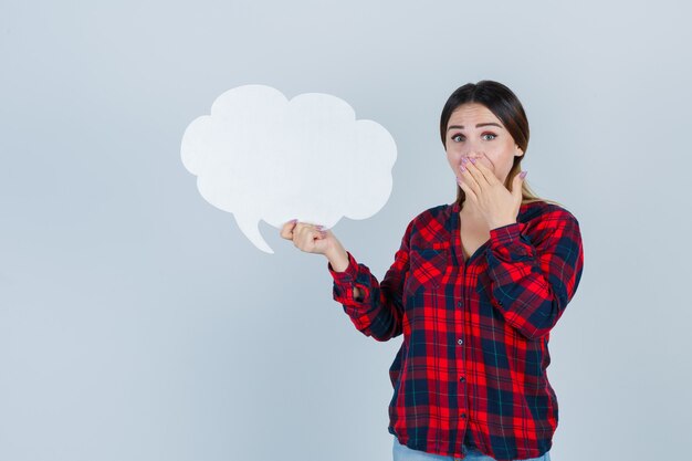 Young beautiful female in casual shirt, jeans covering mouth while holding speech bubble and looking bewildered , front view.