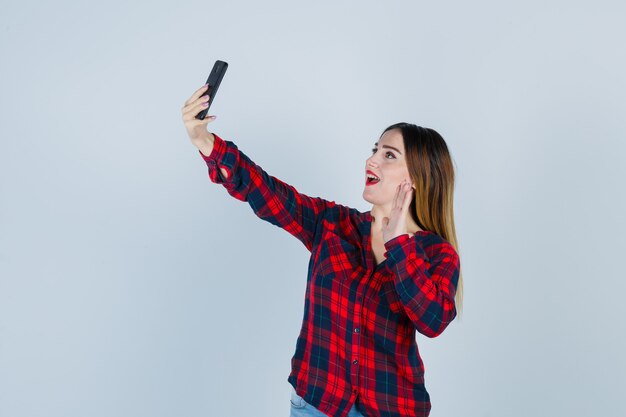 Young beautiful female in casual shirt taking selfie, waving hand and looking joyful , front view.