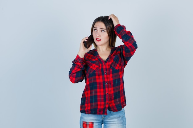 Young beautiful female in casual shirt talking on phone, scratching her head and looking thoughtful , front view.