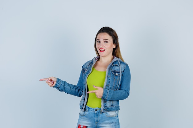 Young beautiful female in denim outfit pointing left and looking cheerful , front view.