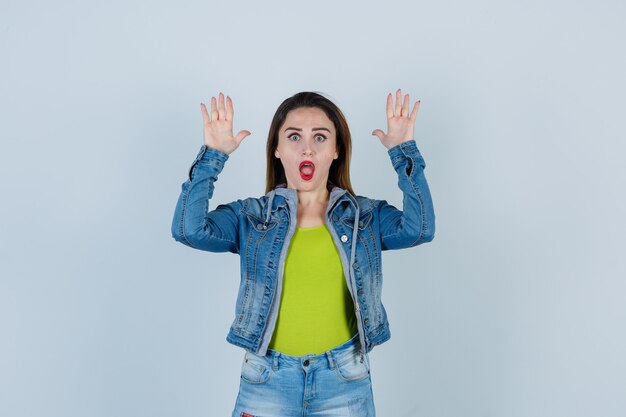 Young beautiful female in denim outfit showing surrender gesture and looking scared , front view.