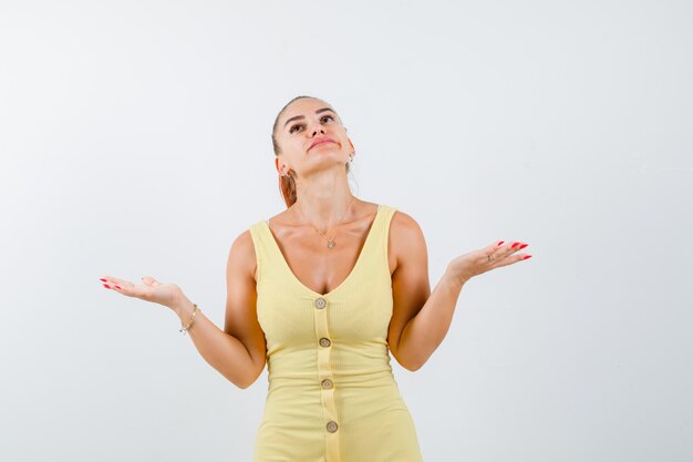 Young beautiful female in dress showing helpless gesture while looking up and looking thoughtful , front view.