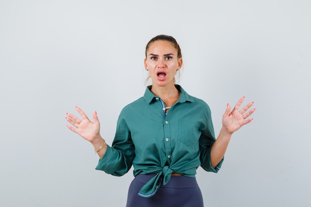 Young beautiful female in green shirt showing surrender gesture and looking scared , front view.