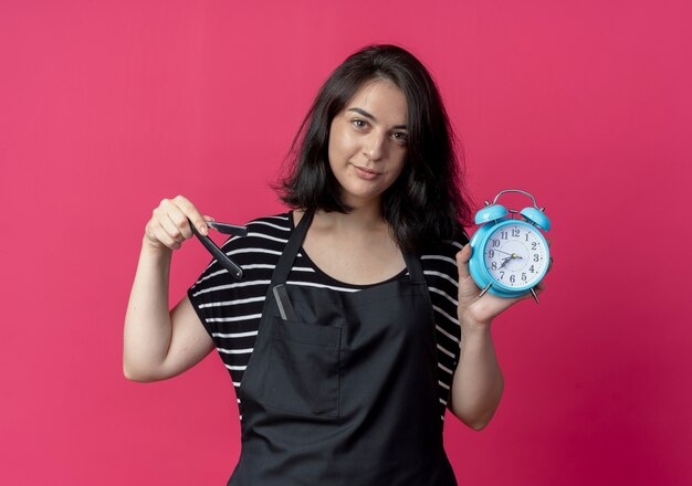 Young beautiful female hairdresser in apron holding alarm clock pointing with finger at it looking confident standing over pink wall