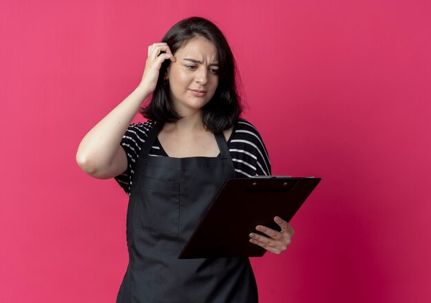 Young beautiful female hairdresser in apron holding clipboard looking at it confused over pink