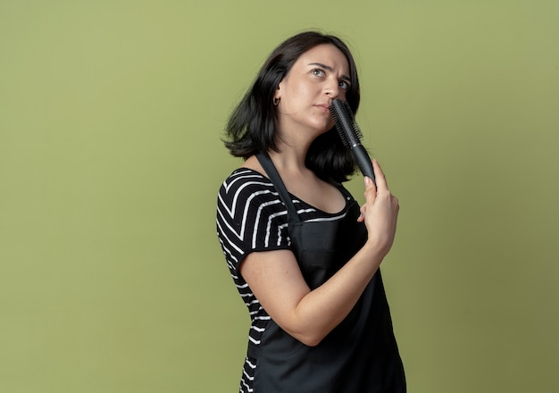 Foto gratuita giovane bella donna parrucchiere in grembiule tenendo le spazzole per capelli guardando da parte in piedi perplesso sopra la parete chiara