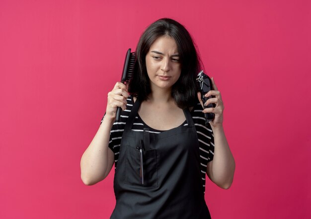 Young beautiful female hairdresser in apron holding hair cutting machine and hair brush looking with serious face  over pink