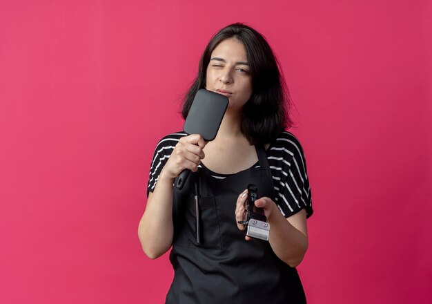 Young beautiful female hairdresser in apron holding hair cutting machine and hair brush smiling and winking  over pink