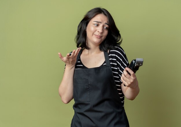 Free photo young beautiful female hairdresser in apron holding hair cutting machine looking at it confused standing over light wall